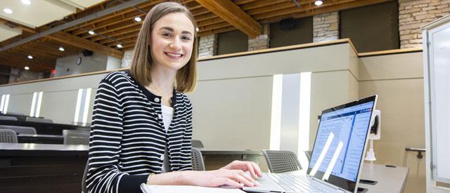 a Carroll University student smiling at the camera and working on a laptop computer.
